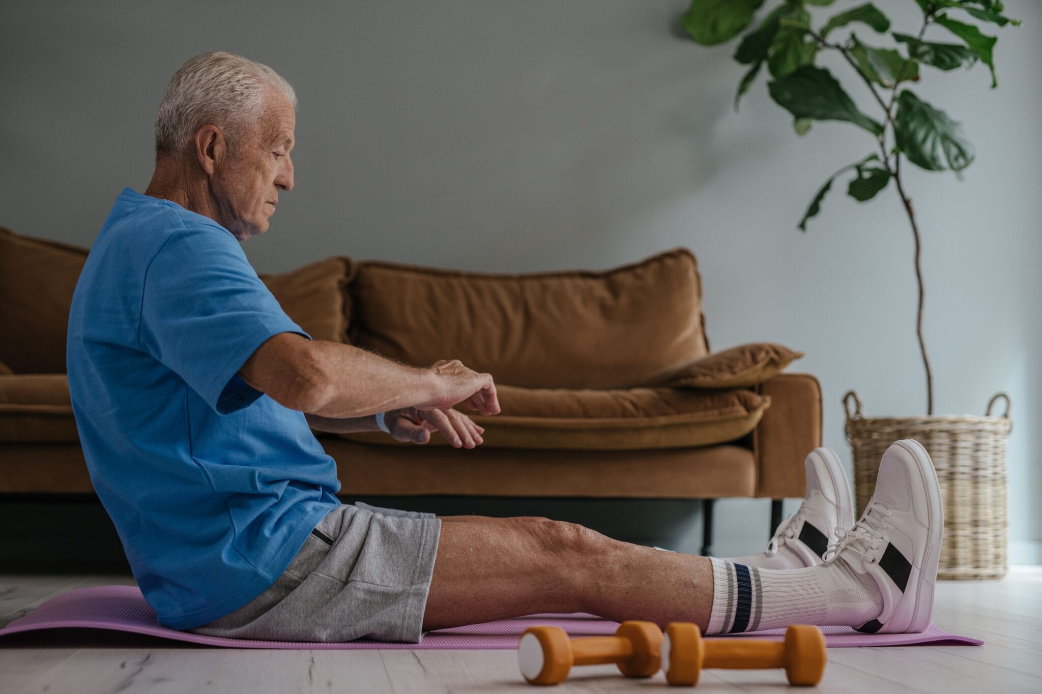 Elderly man in blue shirt exercising on a yoga mat indoors with dumbbells in view.